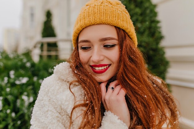 Close-up portrait de charmante femme au gingembre debout près de sapin. Photo extérieure d'une fille souriante joyeuse au chapeau d'hiver.