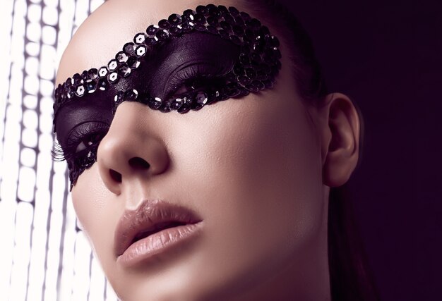 Close-up portrait de charmante femme brune élégante en masque de paillettes posant sur fond noir