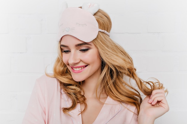 Close-up portrait de femme avec une coiffure glamour posant le matin du week-end. Joyeuse fille blanche dans un masque de sommeil jouant avec des cheveux bouclés.