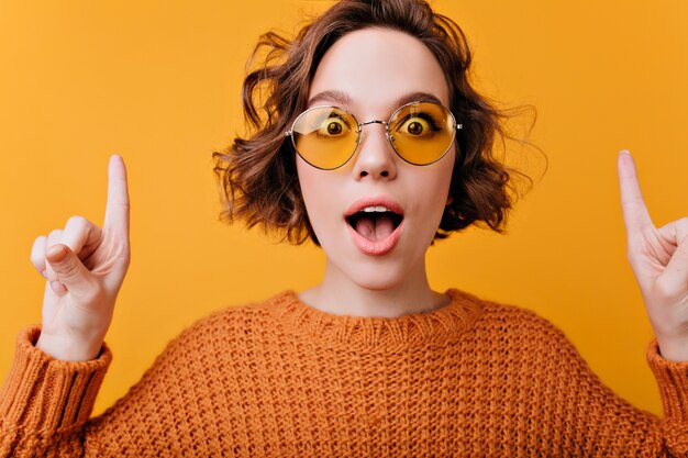 Close-up portrait de femme européenne positive porte d'élégantes lunettes de soleil jaunes. charmante fille bouclée exprimant des émotions surprises.