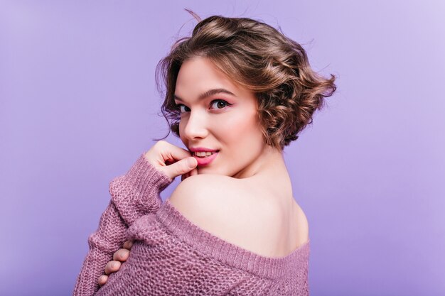 Close-up portrait de jeune femme intéressée aux cheveux courts brune porte un pull. Photo intérieure d'une femme sensuelle regardant par-dessus l'épaule sur un mur violet.