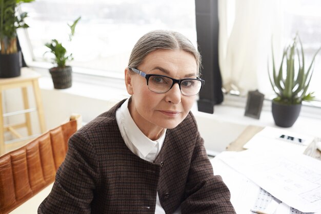 Close up portrait of attractive 60 ans designer femme de race blanche avec des cheveux gris portant des lunettes rectangulaires faire de la paperasse dans son espace de travail léger, à la recherche avec une expression sérieuse