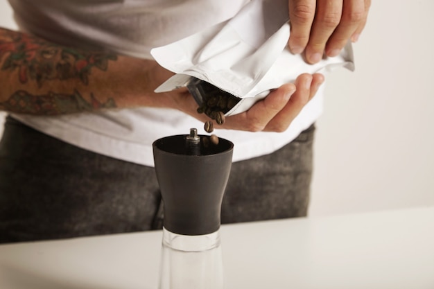 Photo gratuite close up portrait of a barista in white t-shirt et jeans verser des grains de café dans un petit moulin à bavures moderne