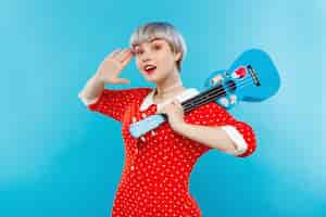 Photo gratuite close up portrait of beautiful dollish girl with short light light hair wearing red dress holding ukulele over blue wall