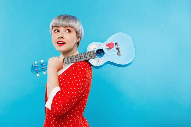 Close up portrait of beautiful dollish girl with short light light hair wearing red dress holding ukulele over blue wall