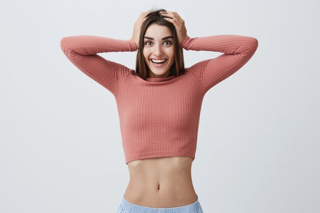 Close up portrait of beautiful joyful young girl with dark long hair in pink elegant top and blue jeans smiling with dents, holding head with hands with happy