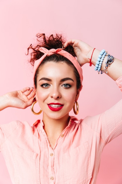 Close-up portrait of blue-eyed lady with red lipstick habillée en chemise rose et boucles d'oreilles massives sur un espace isolé.