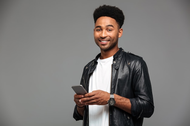 Close-up portrait of cheerful african guy with stylich haircut holding mobile phone, looking