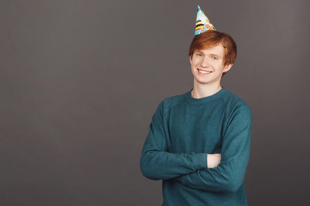 Close up portrait of cheerful male with ginger hair in green sweater and party cap smiling, crossing hands