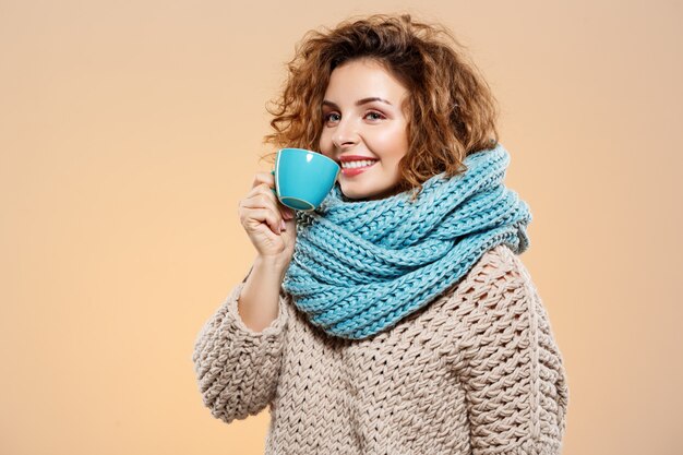 Close up portrait of cheerful smiling beautiful brunette brunette curly girl in knited pull and grey neckwarmer holding cup over beige wall