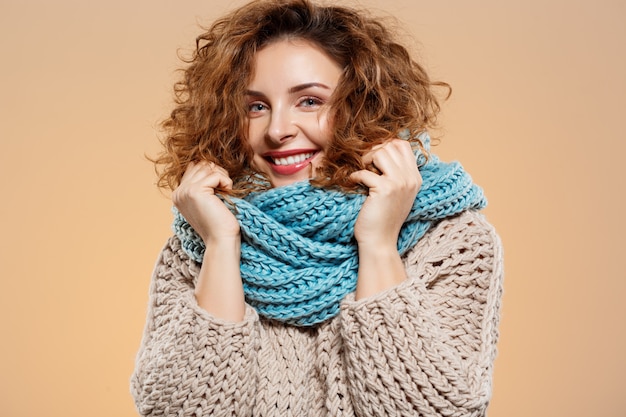 Close up portrait of cheerful smiling beautiful brunette curly girl in pull en tricot et cache-cou gris sur mur beige