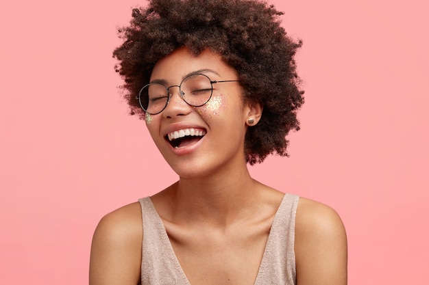 Close up portrait of happy African American female rit de quelque chose de drôle, a une expression positive, porte des lunettes, a les cheveux bouclés, habillé avec désinvolture, ferme les yeux avec bonheur, isolé sur rose