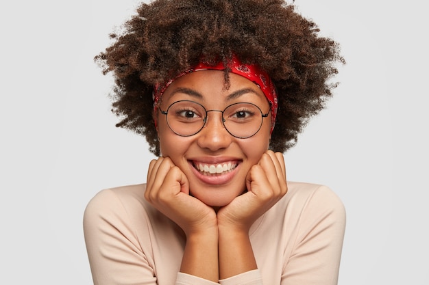 Close up portrait of happy black lady tient le menton à deux mains, heureux que tout va bien, porte des lunettes rondes, a les cheveux bouclés, écoute une histoire drôle de l'interlocuteur. Concept d'émotions positives