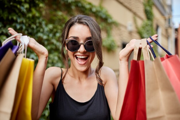 Close up portrait of happy joyful young dark-haired caucasian woman in tab verres et chemise noire regardant à huis clos avec la bouche ouverte et l'expression heureuse, tenant des sacs colorés dans les mains. Gi