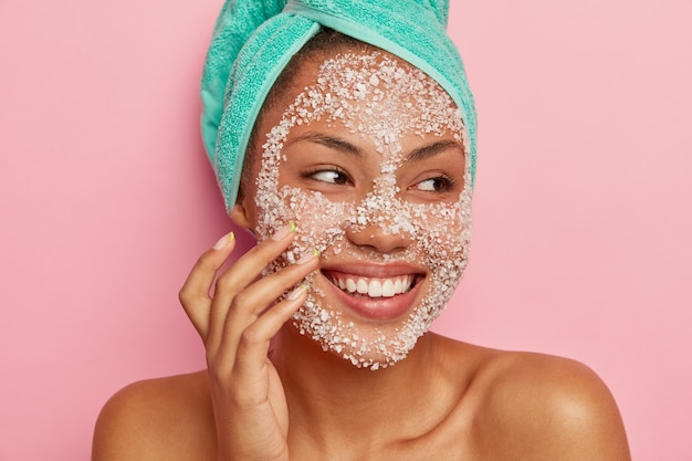 Close up portrait of happy woman massages joues, applique un gommage au sel de mer, regarde ailleurs, a un sourire doux, montre des dents blanches, porte une serviette turquoise, heureux de recevoir des soins de spa.