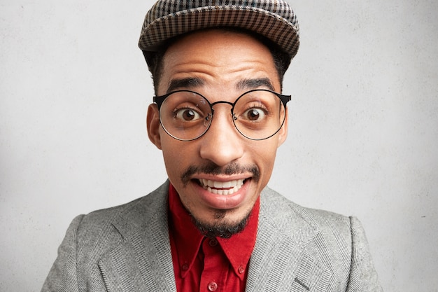 Photo gratuite close up portrait of male comic maladroit porte de grandes lunettes, casquette et veste, sourit de surprise,