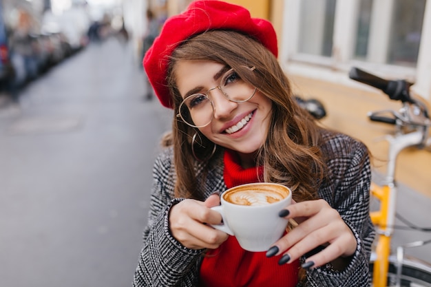 Close-up portrait of pâle fille brune avec charmant sourire tenant doucement la tasse de café