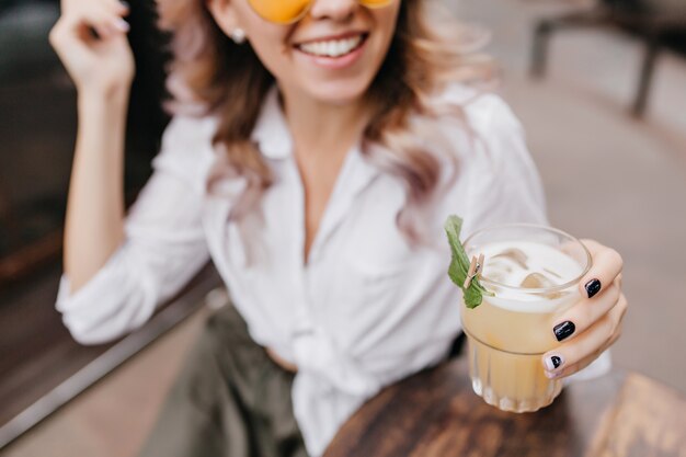 Close-up portrait of smiling lady en chemise blanche avec main tient un verre de café glacé au premier plan