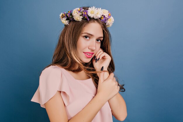 Close-up portrait of timide femme blanche en guirlande de fleurs posant sur mur bleu