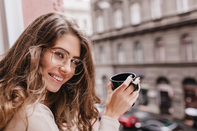 Close-up portrait of wonderful woman holding cup of latte sur fond de ville flou et à la recherche d'appareil photo