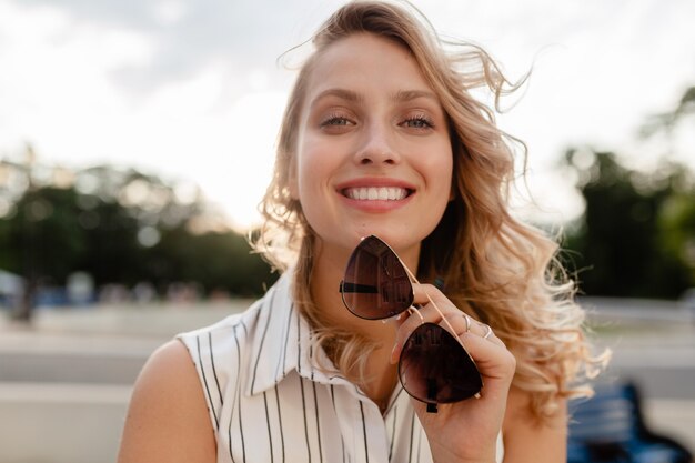 Close-up portrait of young attractive blonde woman in city street in summer fashion style dress holding lunettes de soleil