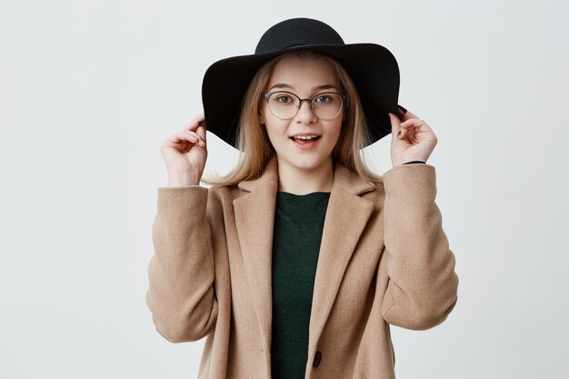 Close-up portrait of young blonde girl with pure skin, eyeglasses and smile wearing black hat and coat isolated. Jolie femme appréciant son style.