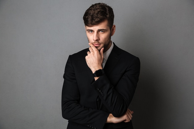 Close-up portrait of young businessman barbu en costume noir tenant son menton,