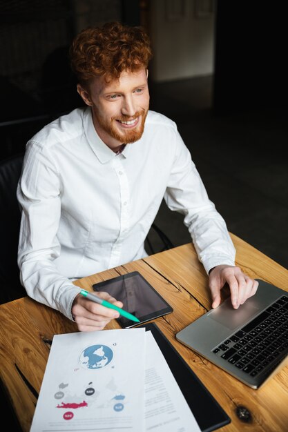 Close-up portrait of young cheerful redhead curly business man in white shirt travaillant sur ordinateur portable, à côté