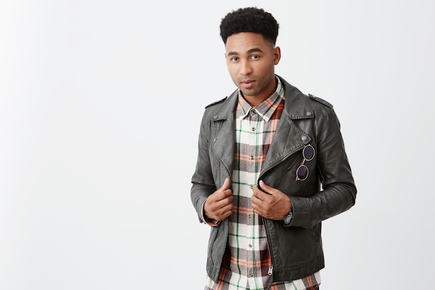 Close up portrait of young handsome dark-skinned american male student with afro hairstyle in leather jacket holding vêtements avec les mains avec une expression confiante et détendue.