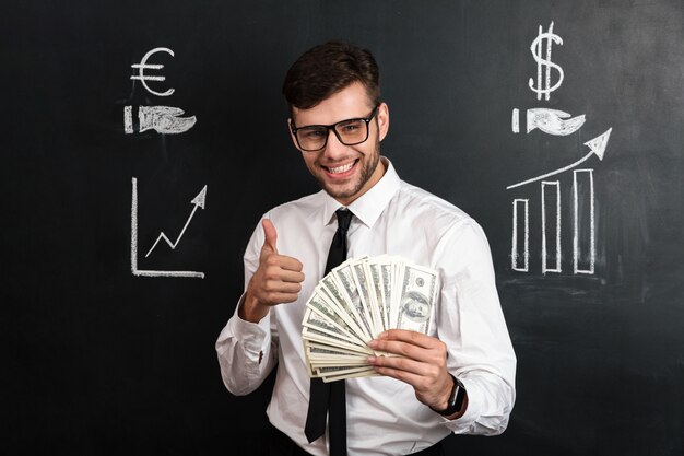 Close-up portrait of young smiling businessman holding bunch of money tout en montrant le pouce vers le haut de geste