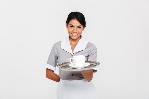 Close-up portrait of young smiling female waiter in uniform holding metal tray with cup of coffee