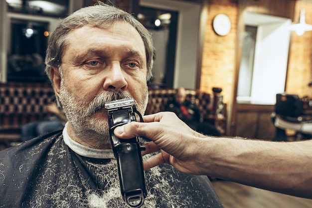 Close-up portrait de vue de côté de bel homme caucasien barbu senior se toilettage de barbe dans un salon de coiffure moderne.