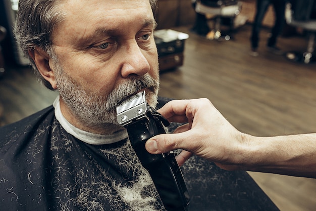 Close-up portrait de vue de côté de bel homme caucasien barbu senior se toilettage de barbe dans un salon de coiffure moderne.