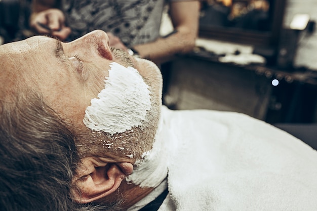 Close-up portrait de vue de côté de bel homme caucasien barbu senior se toilettage de barbe dans un salon de coiffure moderne.