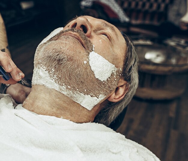 Close-up side vue de dessus beau senior barbu caucasien homme se barbe dans un salon de coiffure moderne.