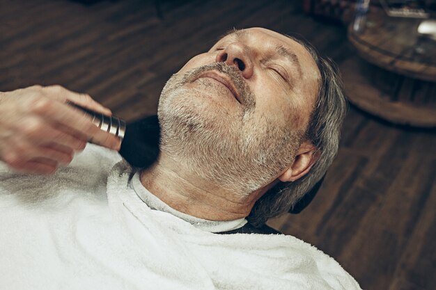 Close-up side vue de dessus beau senior barbu caucasien homme se barbe toilettage dans un salon de coiffure moderne.