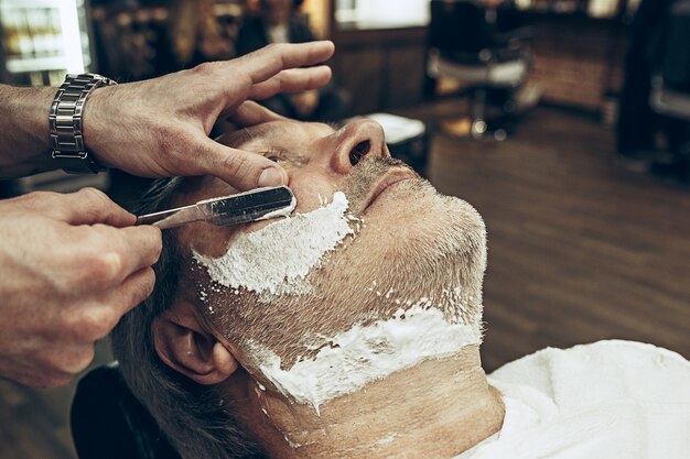 Close-up side vue de dessus beau senior barbu caucasien homme se barbe toilettage dans un salon de coiffure moderne.