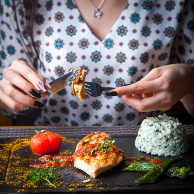 Close-up steak de poulet grillé femme mange avec un couteau et une fourchette avec garniture, tomate, poivre une table en bois sombre horizontal