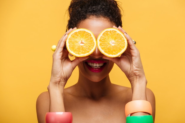 Closeup drôle femme métisse avec des accessoires de mode s'amusant et couvrant les yeux avec deux moitiés d'orange isolé, sur mur jaune