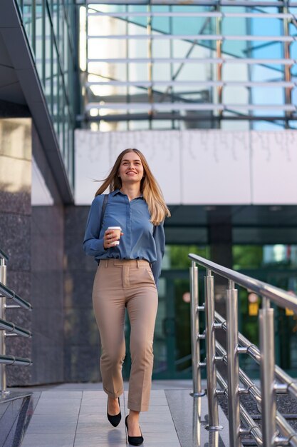 Closeup jolie femme en mouvement avec café à emporter sur le bâtiment de l'entreprise. Portrait fille blonde tenant une tasse de papier avec une boisson chaude en plein air.