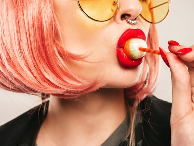 Closeup lèvres de jeune belle hipster mauvaise fille en veste de cuir noir à la mode et boucle d'oreille dans son nez.Sexy insouciante femme souriante posant en studio en perruque rose.Modèle positif léchant des bonbons ronds