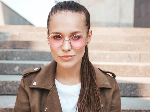 Closeup portrait de beau modèle brune vêtue d'une veste hipster d'été. Fille branchée, assis sur les marches dans le fond de la rue. Femme drôle et positive en lunettes de soleil rondes