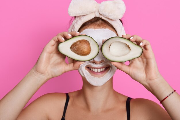 Closeup portrait de femme des années 20 avec une peau parfaite tenant avocat contre ses yeux isolés sur fond rose, soins de santé, procédures cosmétiques à la maison.