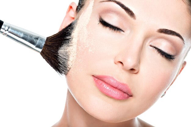 Closeup portrait d'une femme appliquant une fondation tonale cosmétique sèche sur le visage à l'aide d'un pinceau de maquillage.