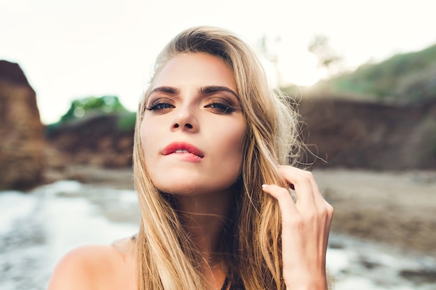 Closeup portrait de fille blonde sexy aux cheveux longs posant sur la plage rocheuse. Elle se mord les lèvres et regarde la caméra.