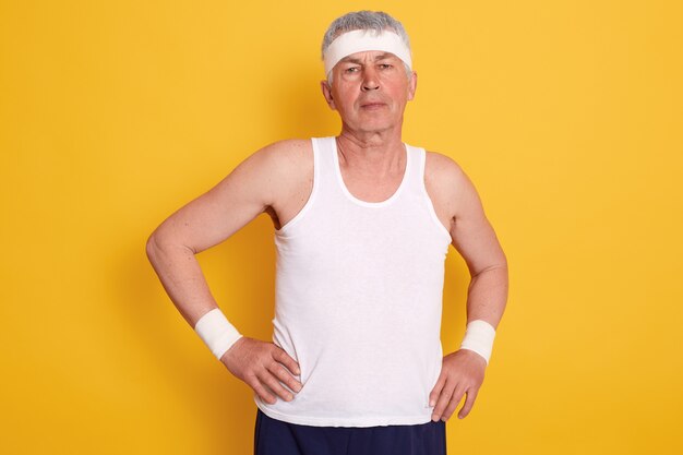 Closeup portrait d'un homme âgé avec les mains sur les hanches, portant un t-shirt sans manches blanc et un bandeau faisant du sport