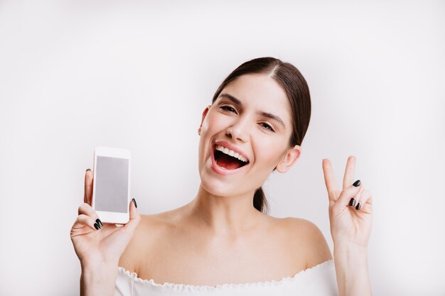 Closeup portrait de jeune fille brune avec téléphone dans ses mains. Jeune femme montre le signe de la paix sur le mur blanc.