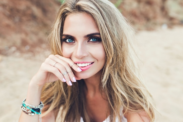 Closeup portrait de jolie fille blonde aux cheveux longs et aux yeux bleus posant sur la plage rocheuse. Elle sourit à la caméra.