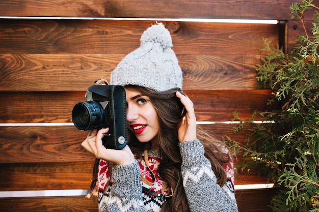 Photo gratuite closeup portrait jolie fille brune en bonnet tricoté et pull chaud faisant une photo à la caméra sur bois. elle sourit.