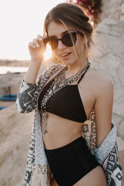 Closeup portrait de jolie jeune femme portant un maillot de bain noir à la mode, bikini, lunettes de soleil élégantes, collier, cardigan, cape avec des ornements sur la plage. Profiter du coucher du soleil, de la station balnéaire, des vacances, des vacances.